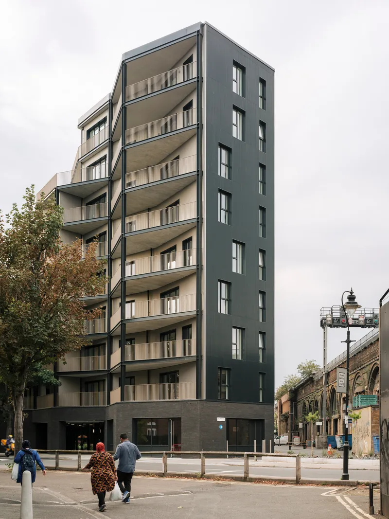 Street view of the eight-storey facade with dark cladding and recessed balconies as two pedestrians cross