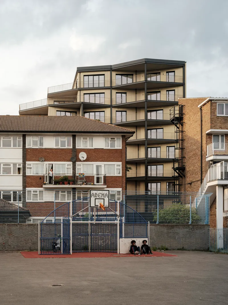 View of the residential building rising behind a brick housing block and basketball court with two seated figures
