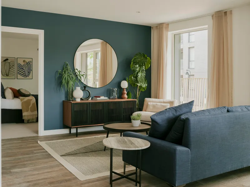 Living room with dark teal accent wall, circular mirror above a credenza, and potted plants by the window
