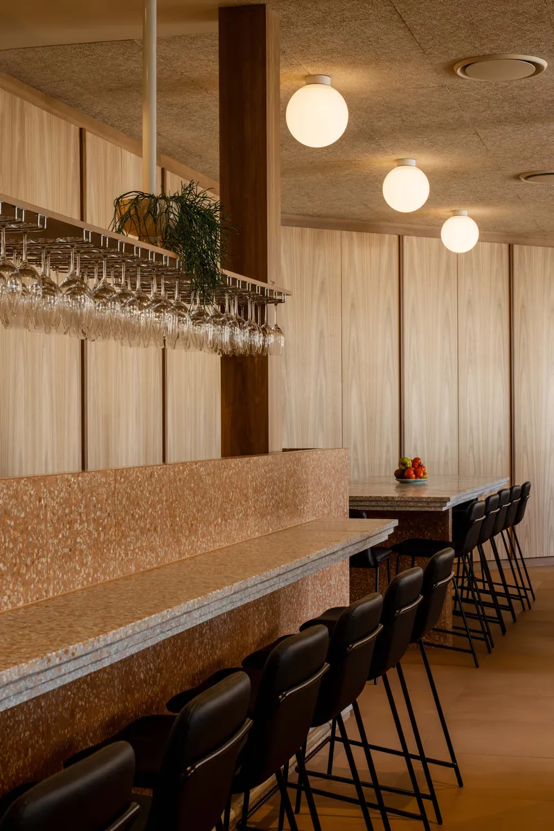 Row of bar stools along terrazzo counter with overhead wine glass rack and globe pendants