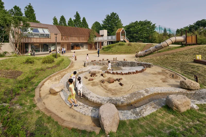 Terraced sand play area with stone edging surrounded by timber-clad pavilions and children at play