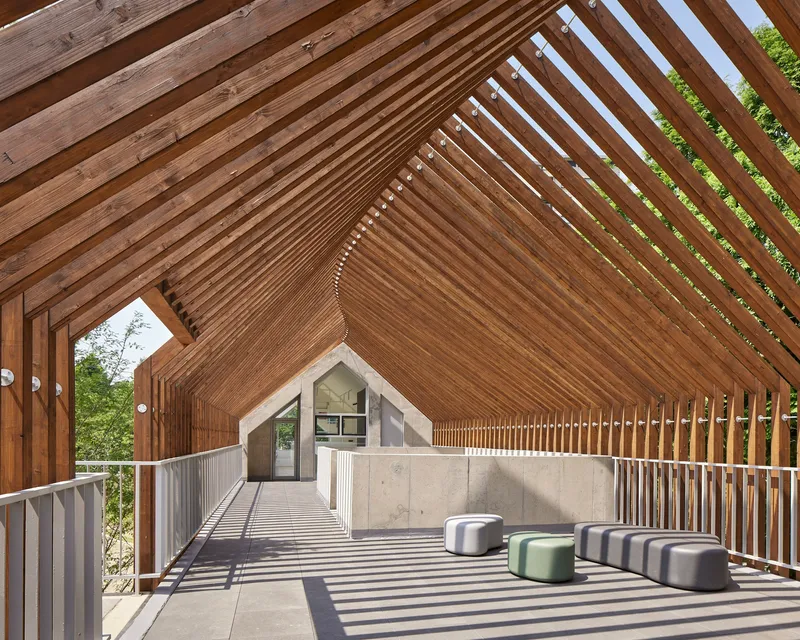 Covered outdoor terrace with slatted timber gable roof filtering afternoon sunlight onto seating pods