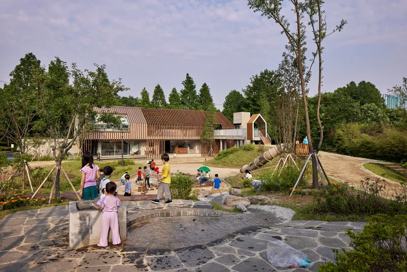 Cluster of buildings with corrugated roofs and timber screens around a landscaped courtyard where children play