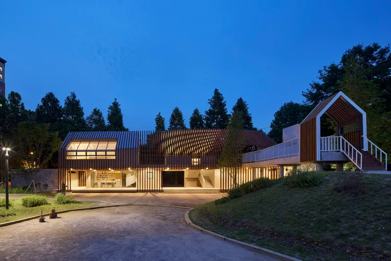Full length view of the timber-clad volumes with illuminated interiors and sloped lawn at dusk