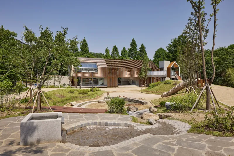 Curved stone paving and shallow water channel leading toward the timber-clad volumes under clear sky