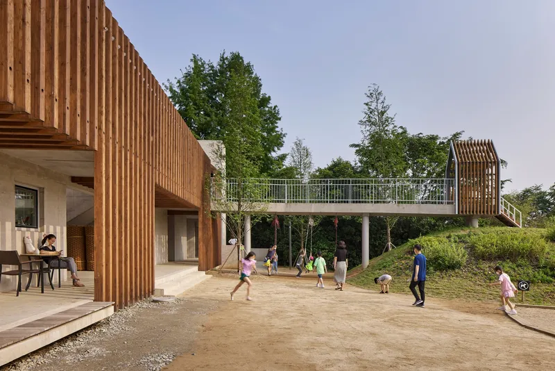 Elevated timber-slatted bridge connecting two volumes above a courtyard with children playing
