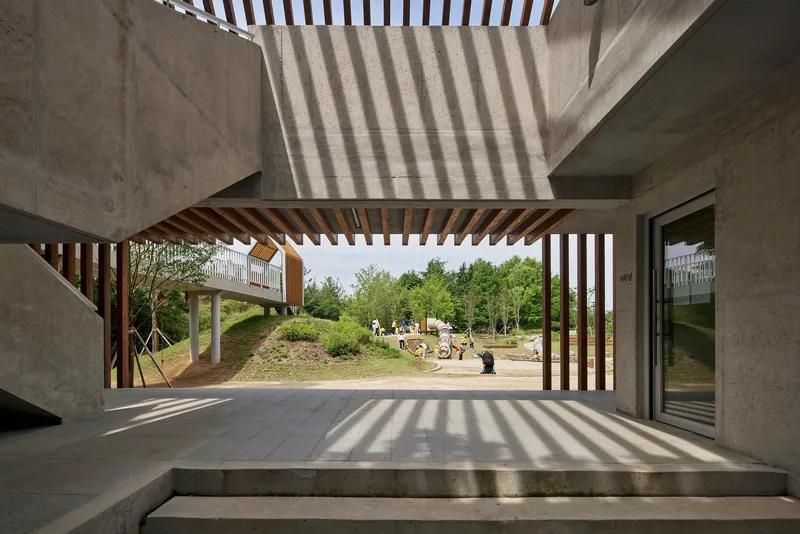 View through timber-slatted porch framing playground and wooded landscape in afternoon light