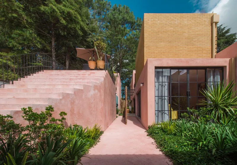 Pink stucco pathway winding past gridded glazing and a yellow brick volume among tropical planting