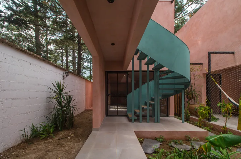 Teal spiral staircase in a courtyard with pink stucco walls and planted beds beneath pine trees