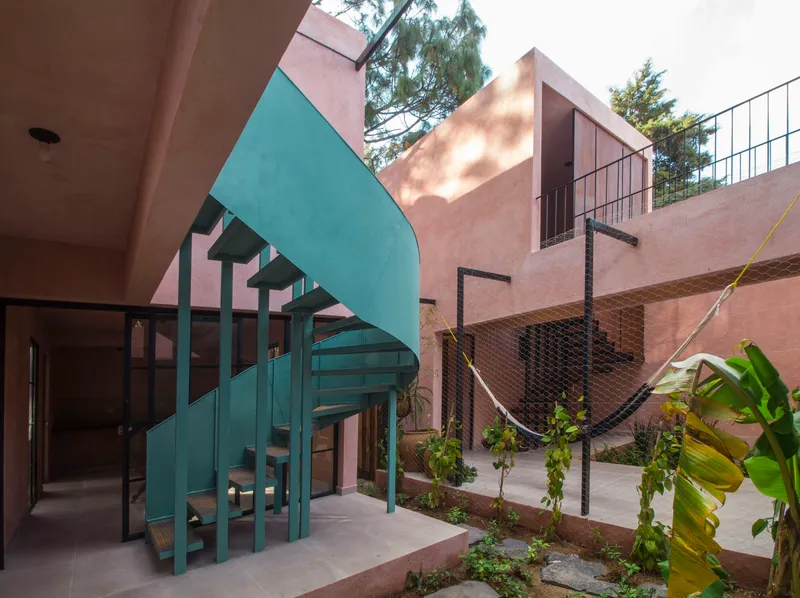 Courtyard view showing the teal spiral stair, hammock netting, and pink stucco volumes in dappled sunlight