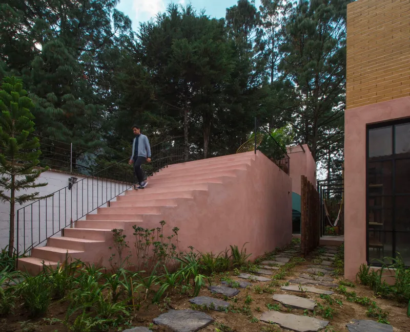 Exterior pink stucco staircase with metal railing and person ascending, surrounded by stone pavers and vegetation