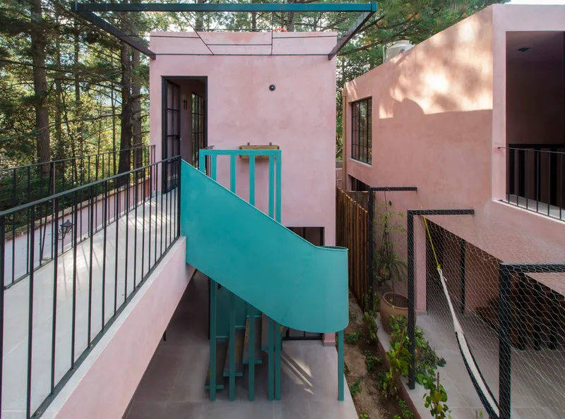 Overhead walkway with metal railing looking down at the teal spiral staircase between pink stucco volumes