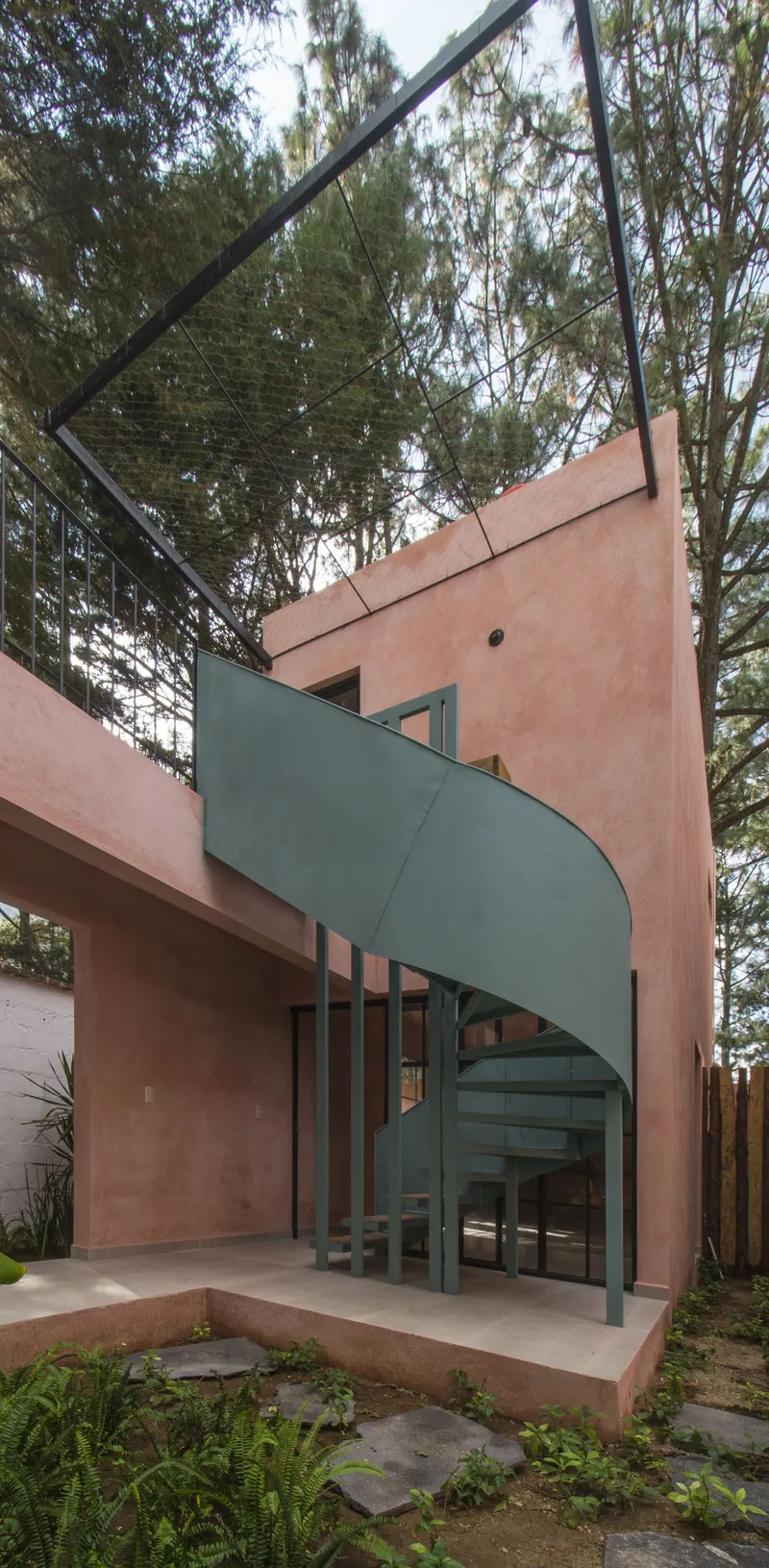 Curved green steel staircase ascending between pink stucco walls beneath tall pine trees
