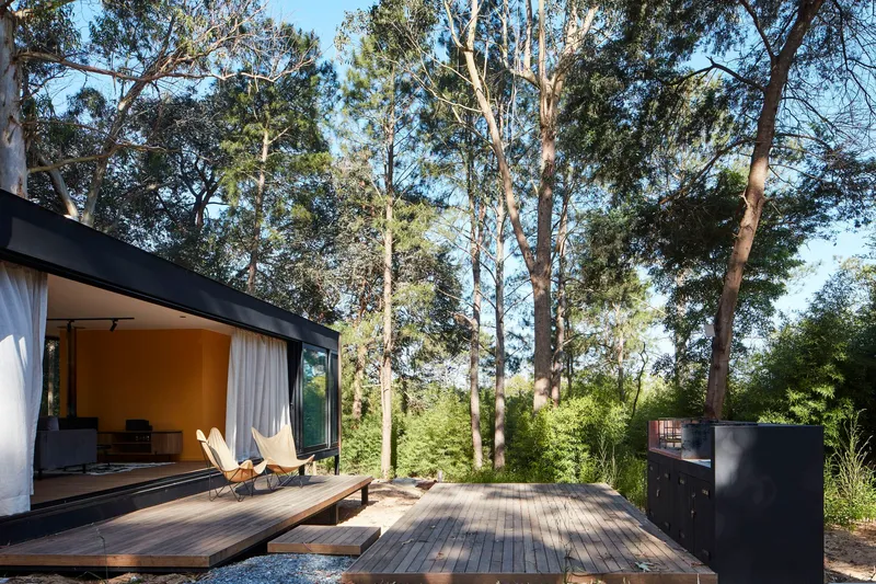 Timber deck terrace extending from the glazed living room toward a freestanding black volume in the trees
