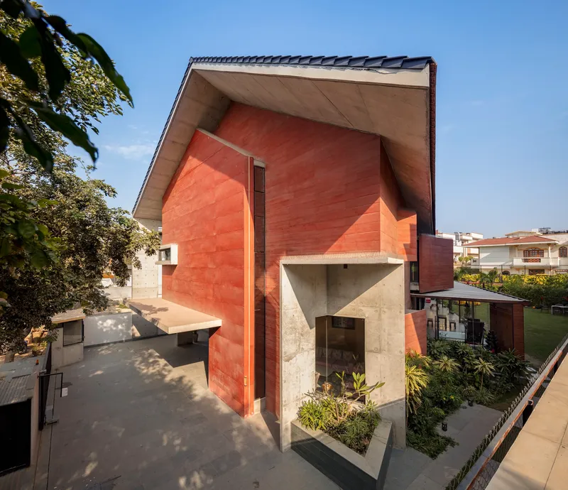 Red volcanic stone facade with angular concrete entry canopy and planted beds under a blue sky