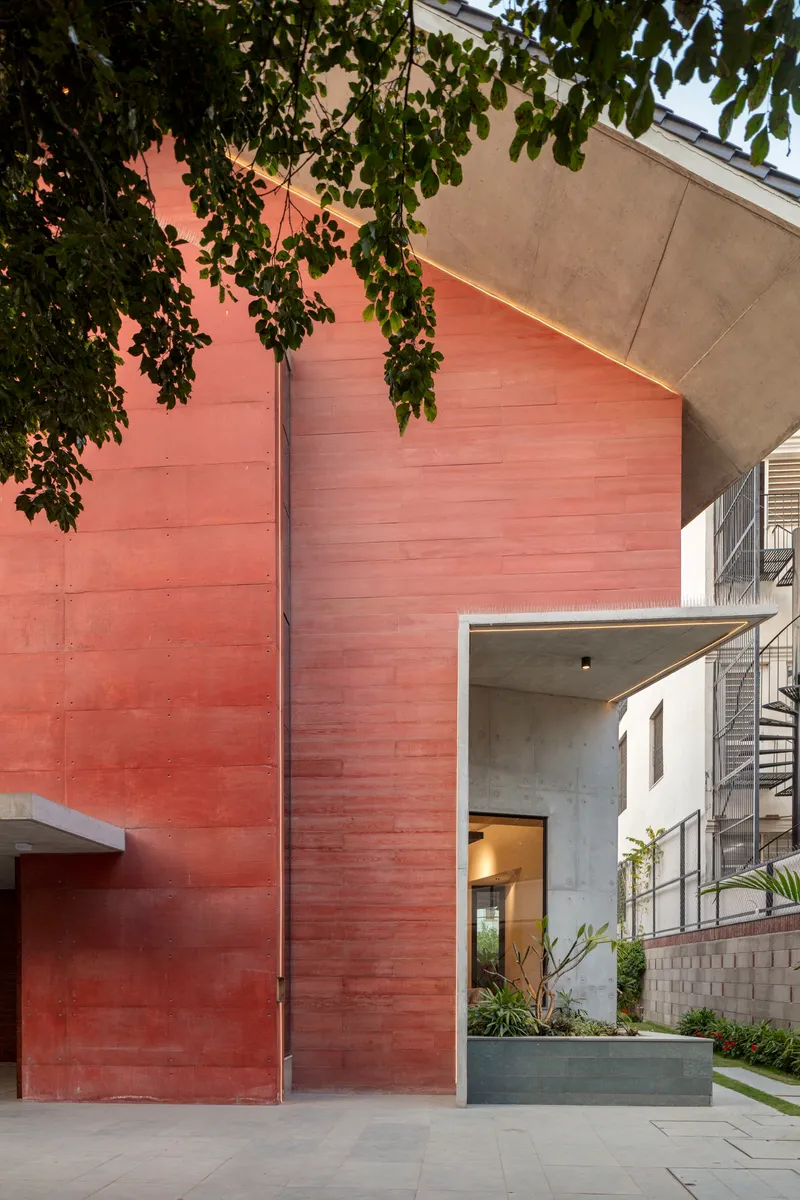 Red stone wall surface meeting concrete entrance volume framed by overhanging tree branches at dusk
