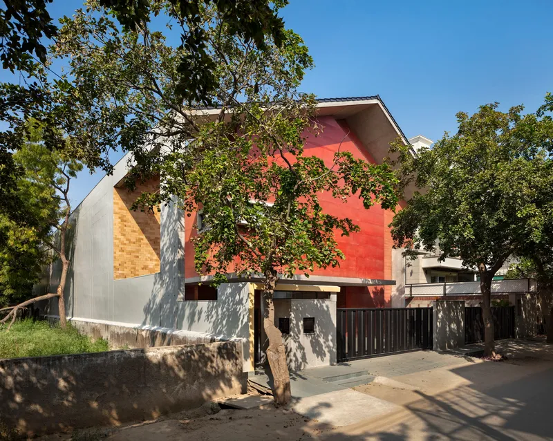 Street view of the gabled form with red-painted facade and diagonal brick cladding framed by mature trees