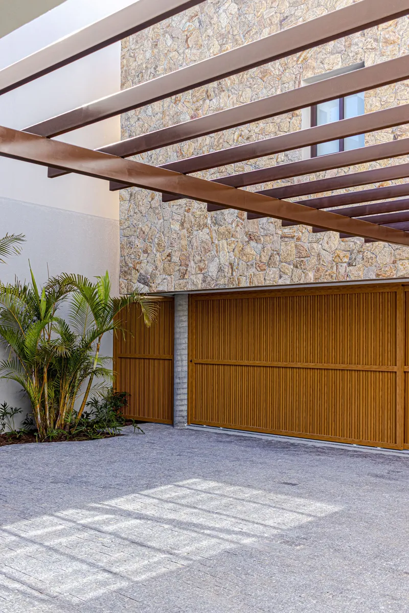 Carport with slatted timber overhead beams and garage door against a stone-clad facade with palms in the foreground