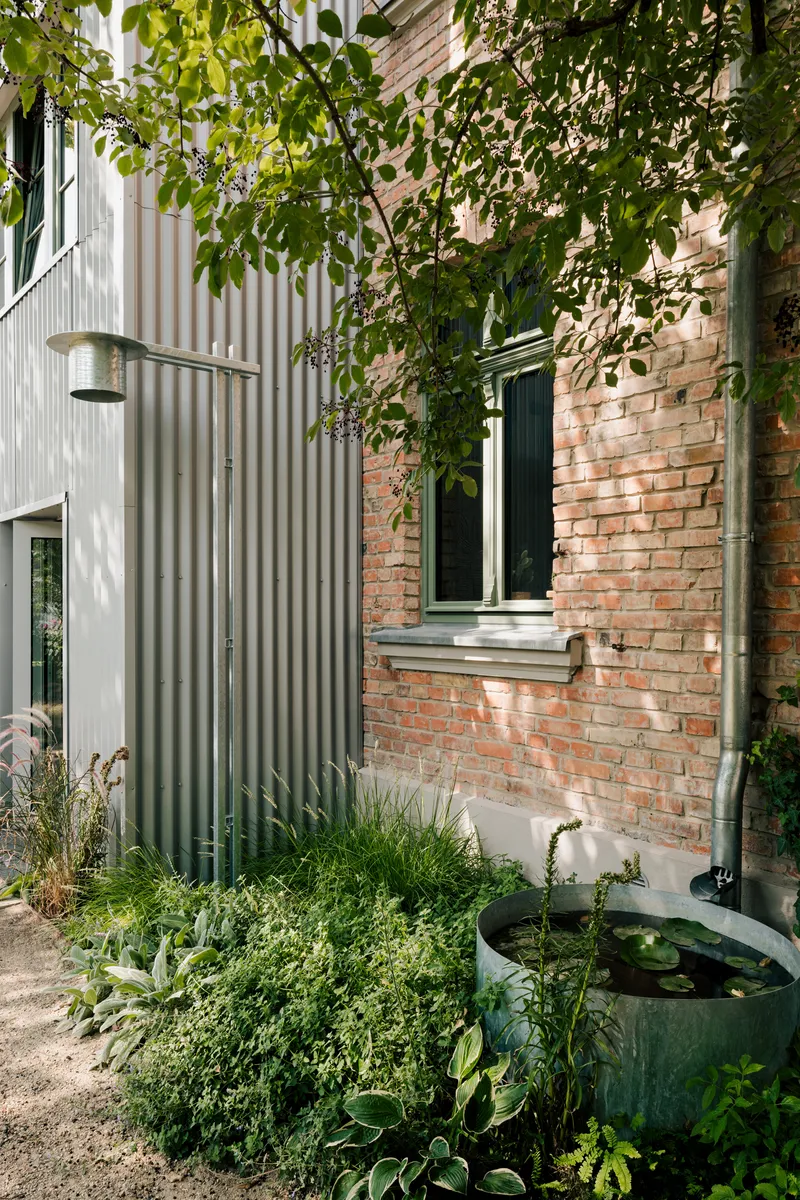 Corrugated metal extension adjoining weathered brick wall with planted garden beds and water basin