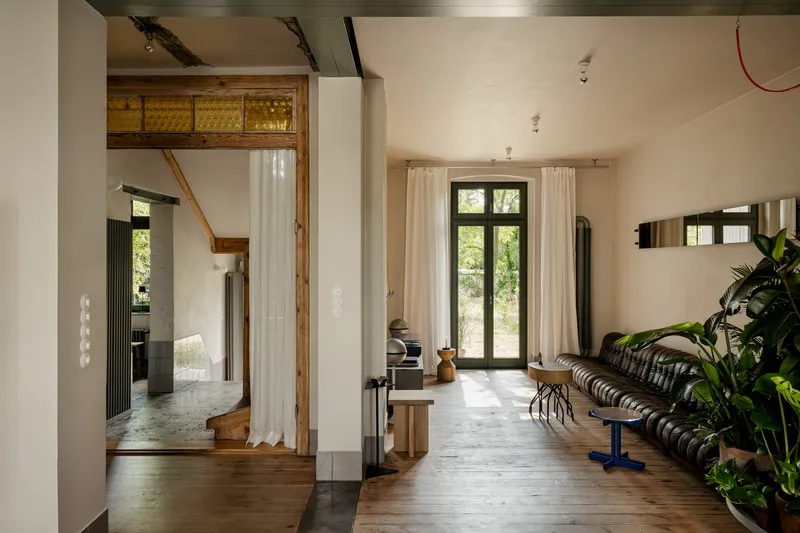 Open interior with exposed timber framing, white curtains, and tall black-framed doors leading to greenery outside