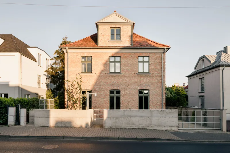 Three-story brick facade with orange tile roof and concrete wall at the street edge in evening light
