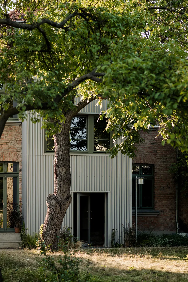 Corrugated metal facade addition with large tree in foreground and aged brick walls at dusk