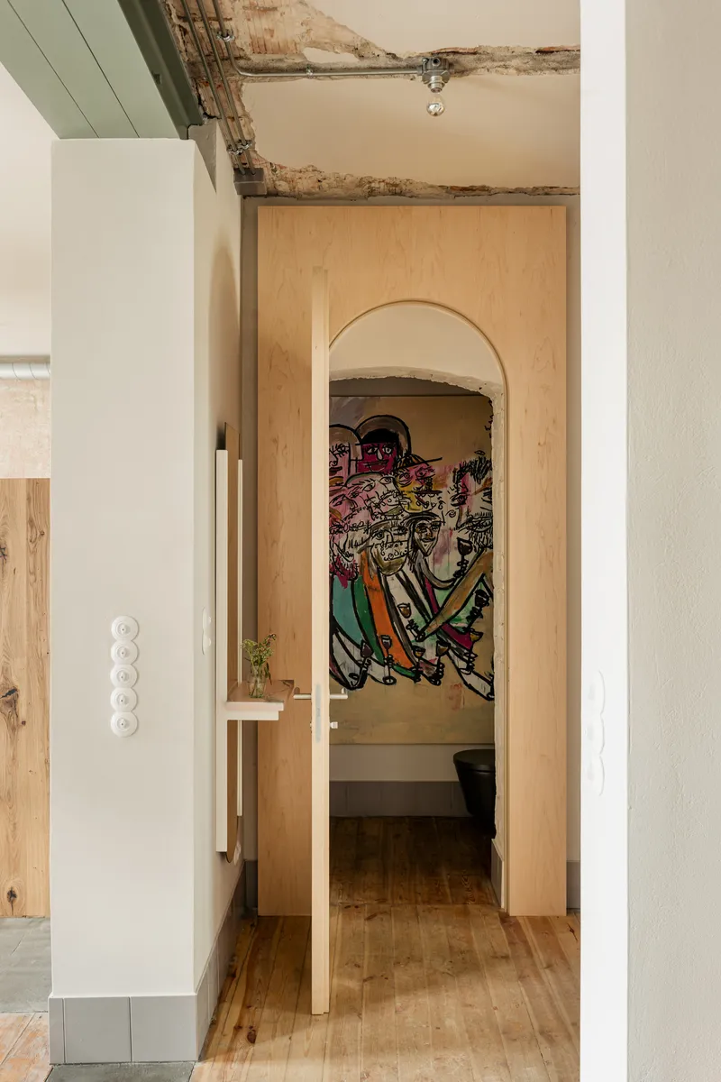 Arched plywood doorway revealing a bathroom alcove with exposed ceiling beams and colorful mural