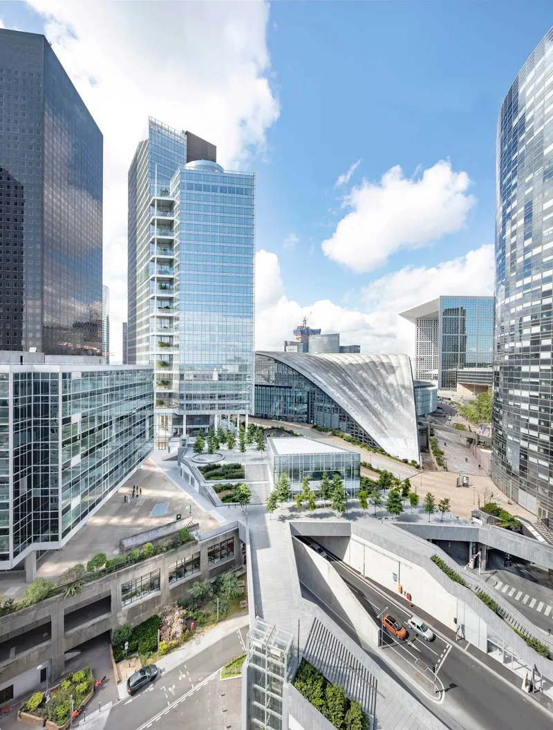 Plaza level with planted terraces and curved shell roof between glass towers on a sunny day