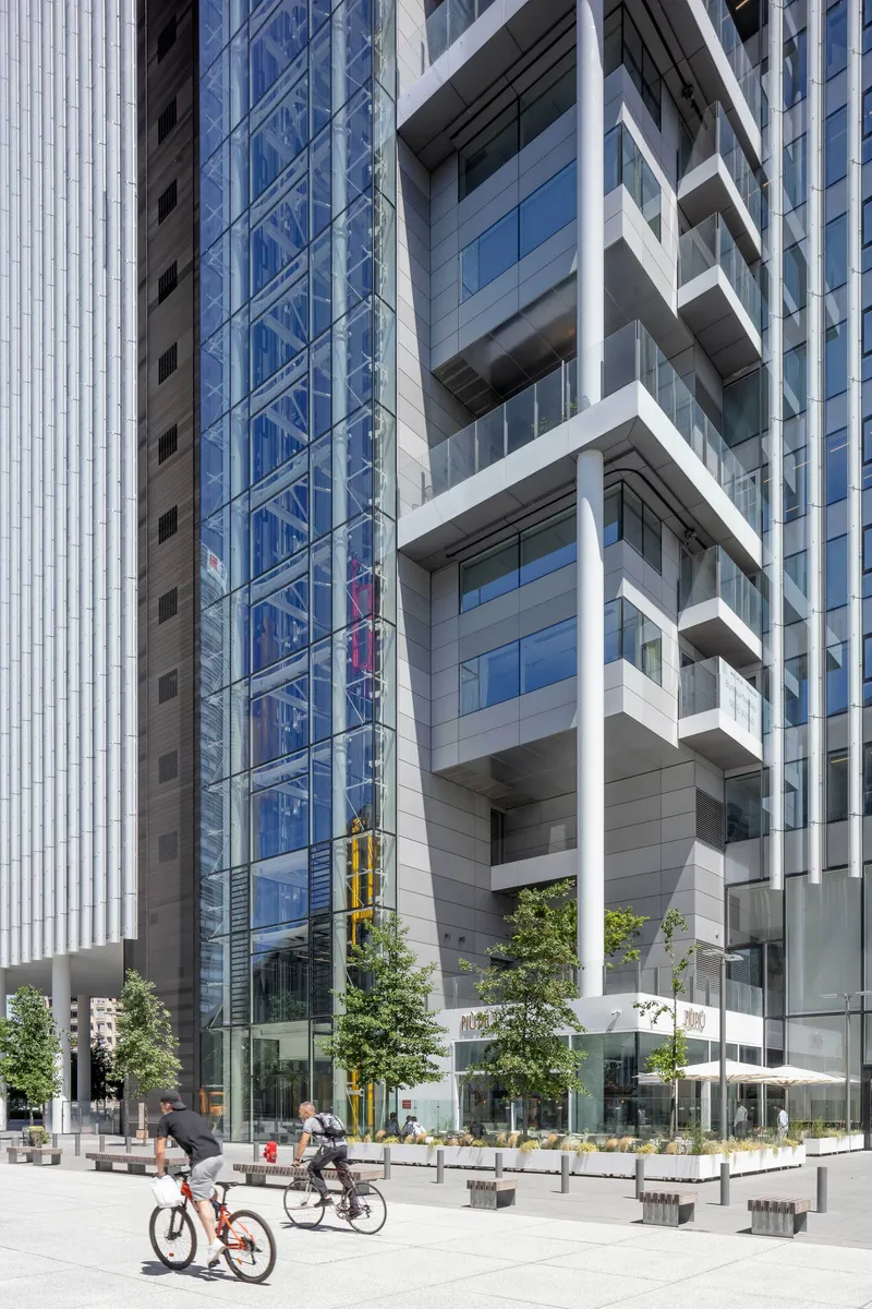 Street view showing angled white balconies on the tower facade with cyclists and trees below