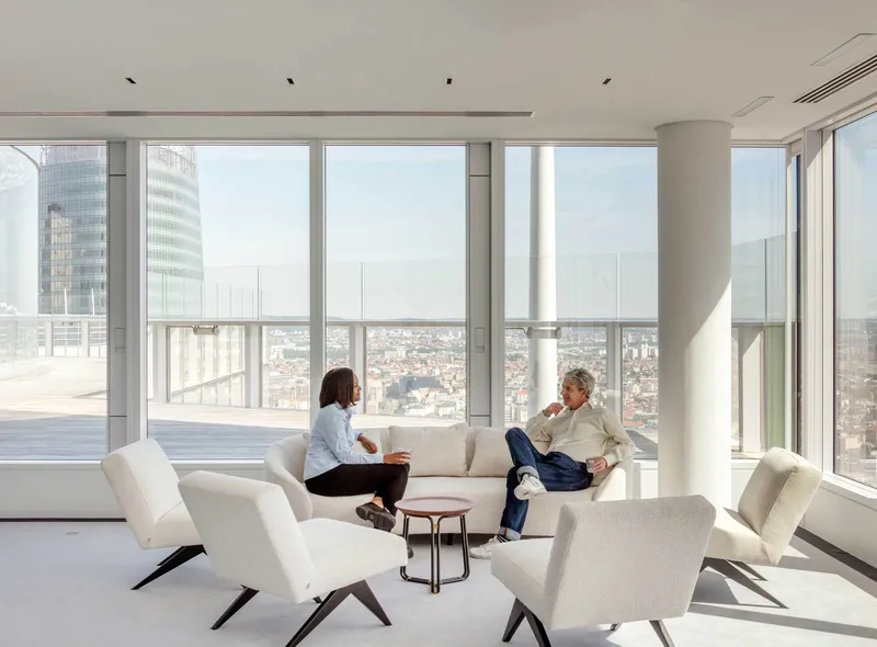 High-rise lounge with floor-to-ceiling glass, white upholstered seating and two people conversing overlooking the city