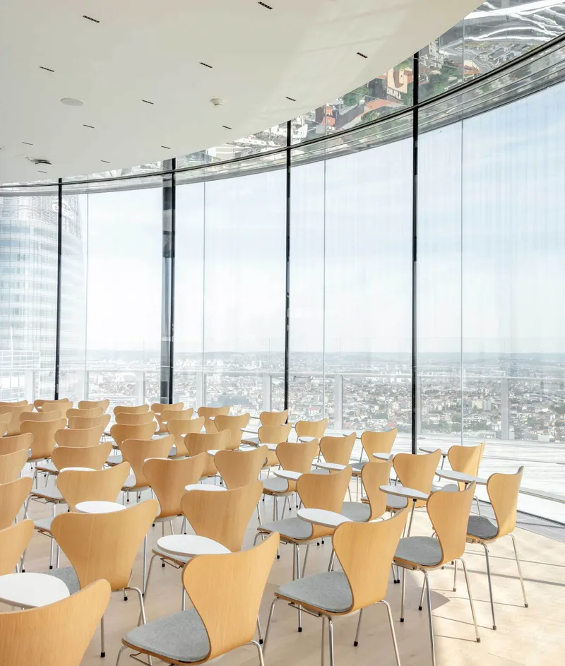 Conference room with rows of plywood chairs facing curved glass walls and panoramic views in bright daylight