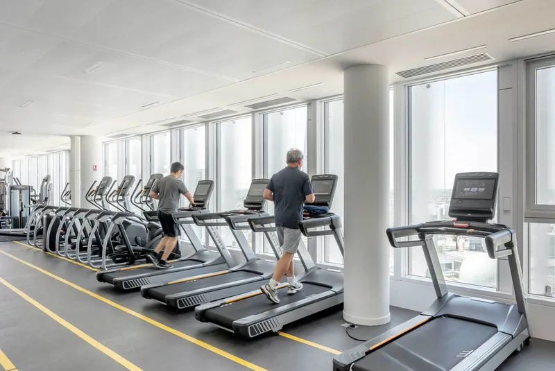 Fitness center with treadmills along the window wall and two people exercising with city views beyond