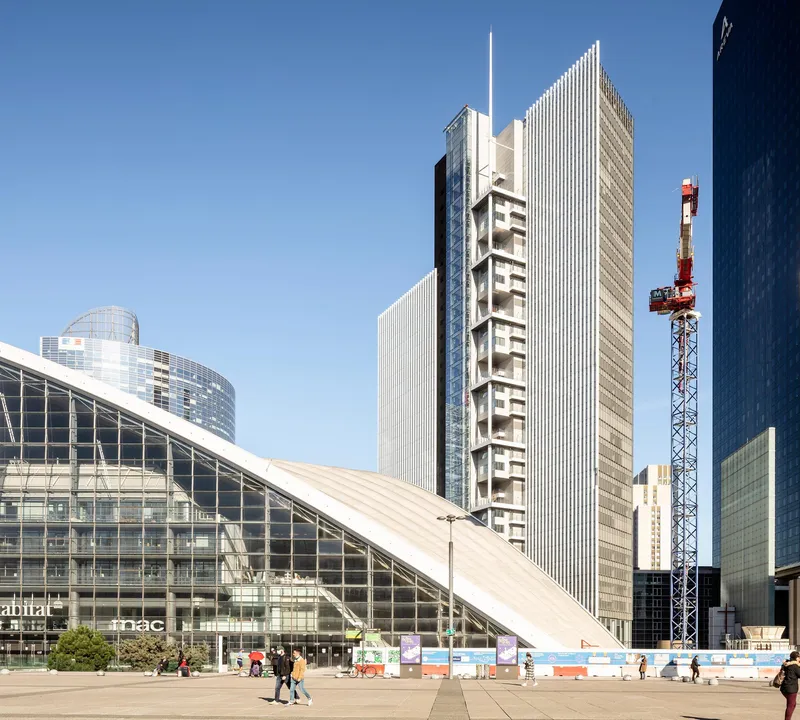 Tower with vertical ribbed facade and central column of balconies seen across a public plaza with construction crane