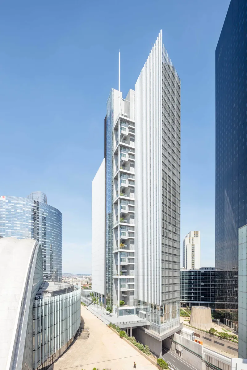 White louvered facade rising above a planted roof terrace with pedestrians and neighboring glass buildings