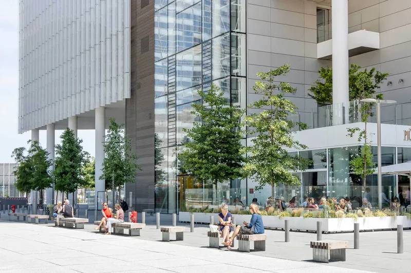 Ground-level plaza with white planters, young trees and people seated on benches beside glass facades