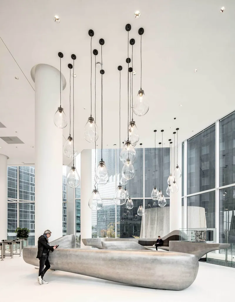 Double-height lobby with clustered pendant lights hanging above curved concrete seating and a visitor reading