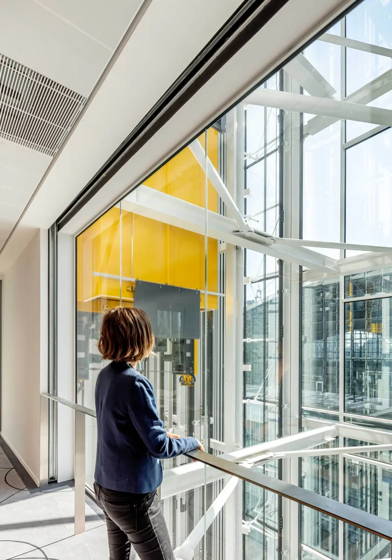 Person standing at a glass balustrade overlooking a multi-story atrium with yellow accent panels