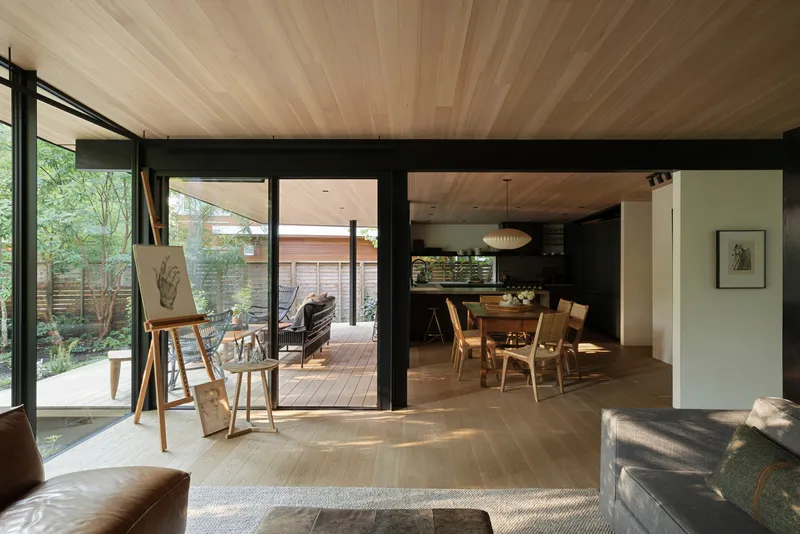 Open-plan living and dining space with black steel framing and radial wood ceiling under dappled sunlight