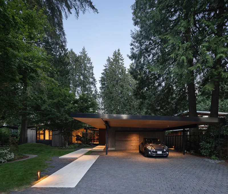 Cantilevered roof over a carport and entrance path surrounded by tall evergreen trees at dusk