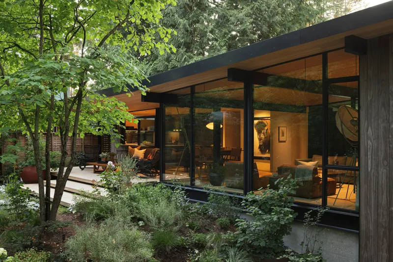 Glass-walled living pavilion framed by planted beds and deciduous trees in evening light
