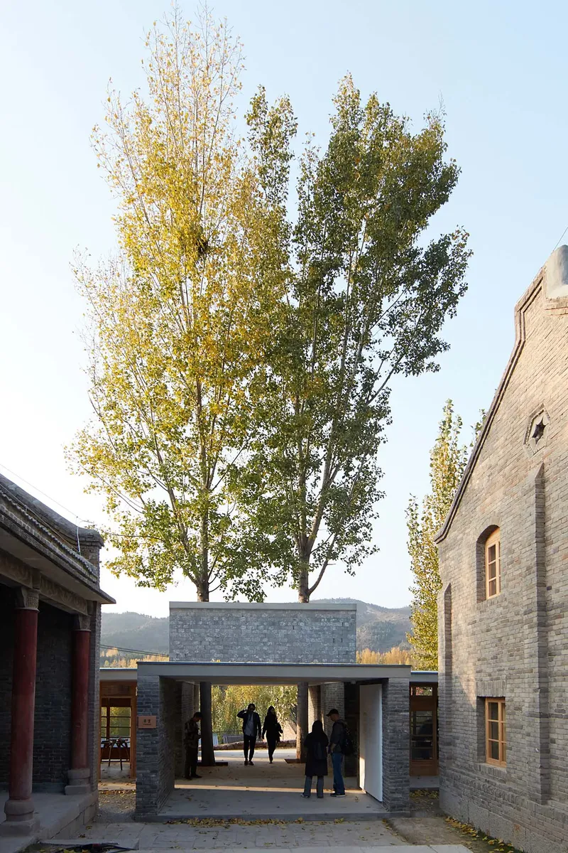 Grey brick pavilion with flat roof connecting two existing structures under tall trees in autumn