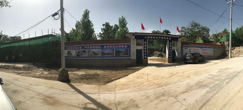 Wide-angle street view showing compound entrance with green fence and red flags under a cloudy sky