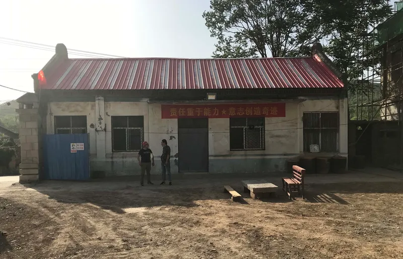 Front facade with corrugated metal roof and two figures standing in the dirt courtyard in haze