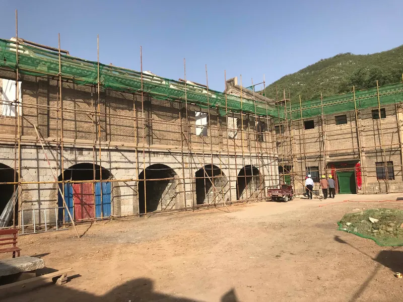 Renovation site showing arched openings wrapped in bamboo scaffolding and green mesh netting with hillside beyond