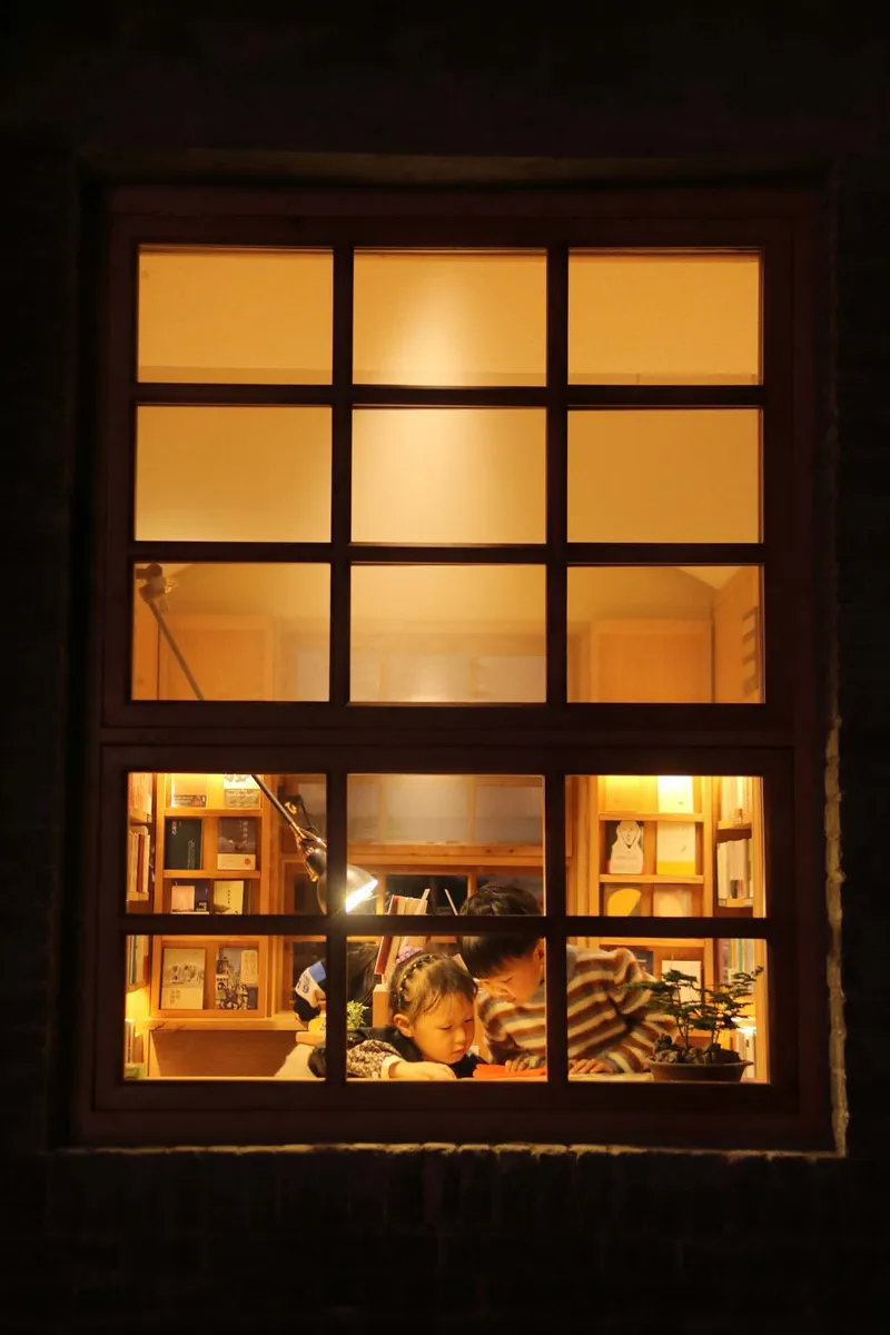 Exterior night view of a gridded window revealing two figures reading inside a warmly lit interior