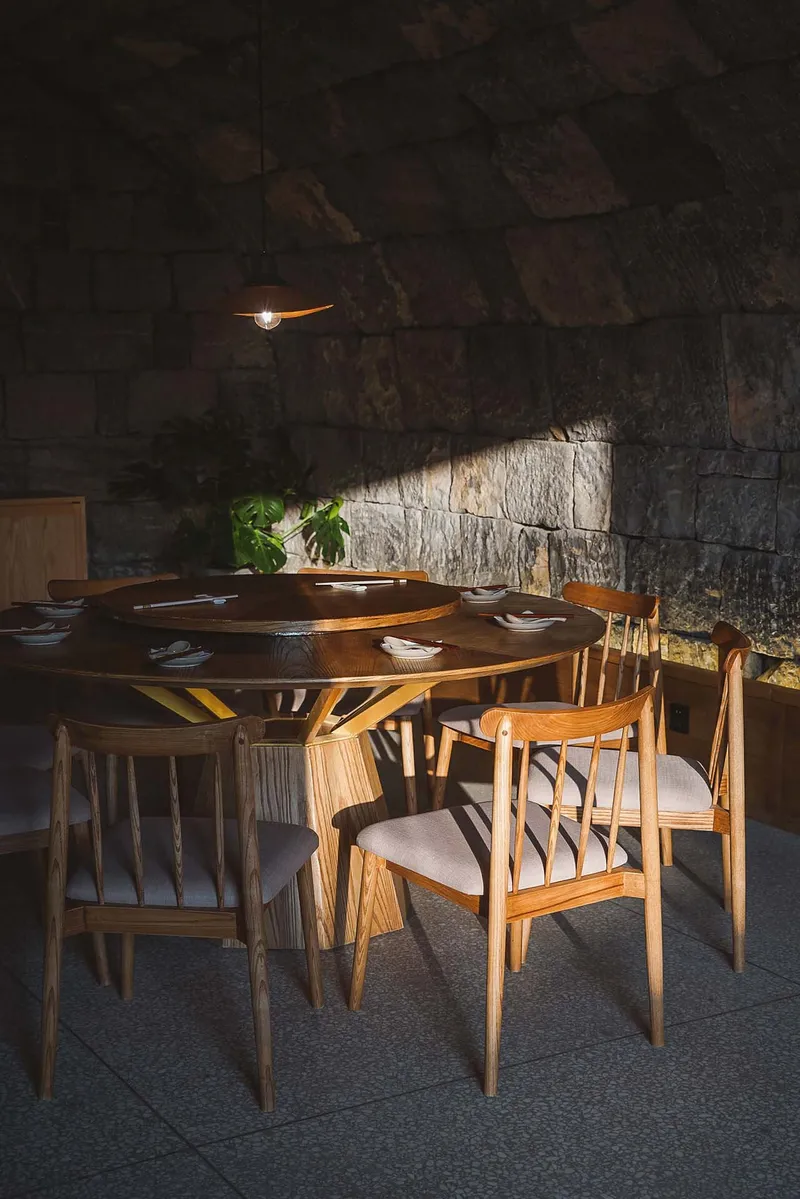 Dining area with a circular timber table surrounded by chairs beneath a pendant light and rough stone wall
