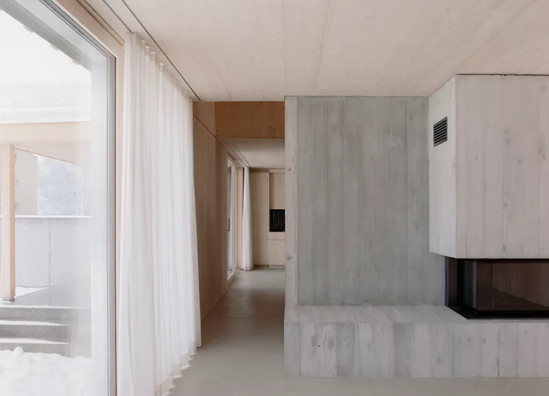 Interior corridor with exposed concrete fireplace mass, plywood ceiling, and floor-to-ceiling sheer curtains along glazing
