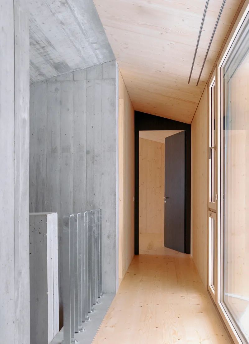 Narrow hallway with board-formed concrete wall, vertical radiator, and plywood-lined ceiling meeting a dark doorway