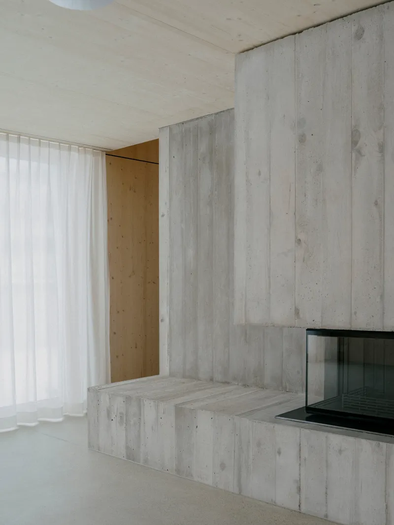 Board-formed concrete fireplace bench and wall beside a translucent curtain and plywood ceiling