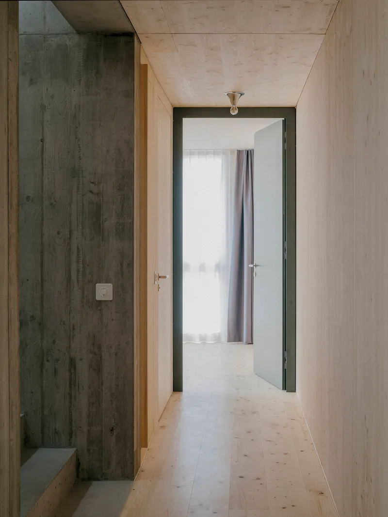 Narrow hallway with light plywood and dark timber walls leading to a room with sheer curtains